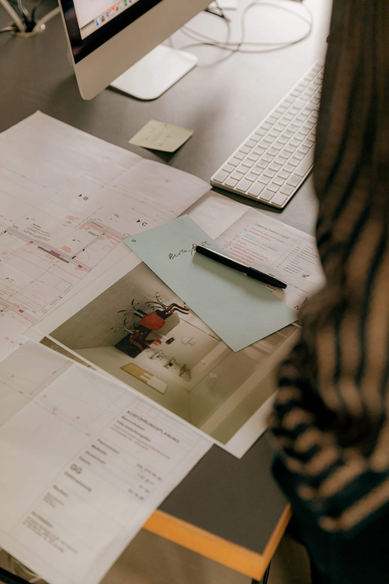 Plans d'étage, photo d'intérieur imprimée et stylo étalés sur un bureau près d'un clavier, vus par-dessus l'épaule d'une personne