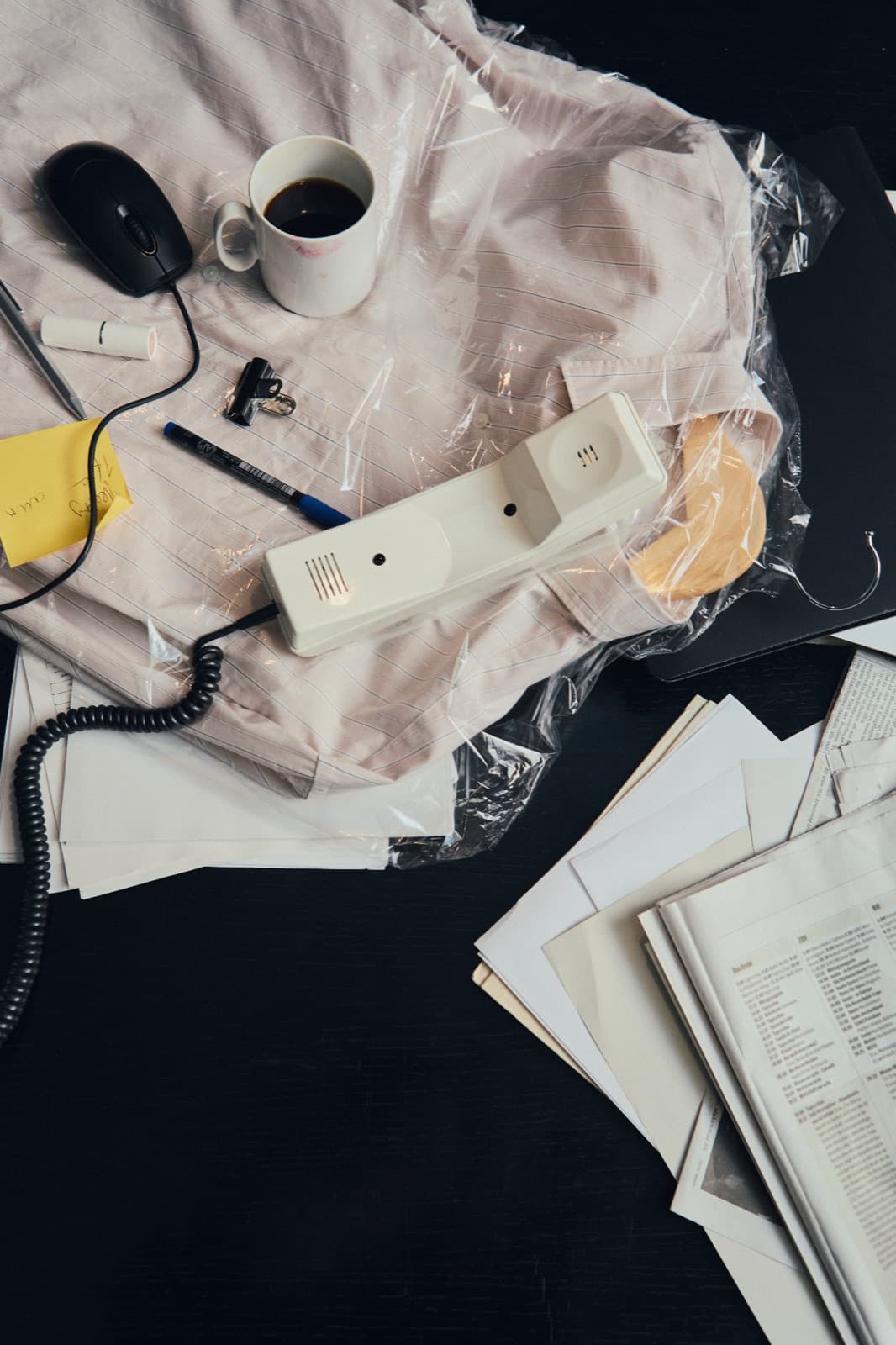Bureau encombré vu de haut : combiné filaire, mug de café et souris sur une chemise emballée sous plastique, près de papiers épars